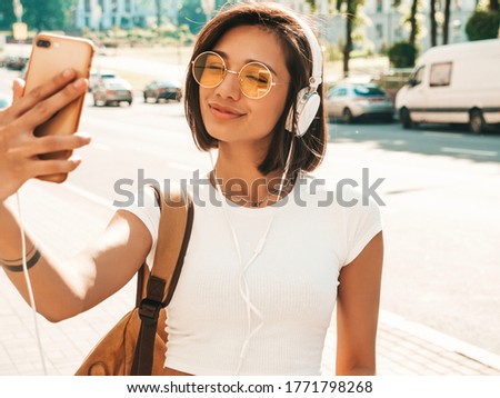 Fashion portrait of young stylish hipster woman walking in the street.Girl making selfie.Smiling model enjoy her weekends with backpack. Female listening to music via headphones