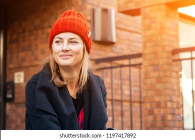 Fashion portrait of young trendy woman dressed in black coat and  red knitted hat sits on bench, look at the camera, enjoy vacation  in the  old city, autumn street fashion.  portrait of joyful woman