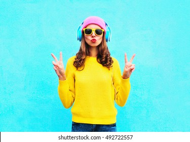 Fashion Pretty Cool Girl In Headphones Listening To Music Wearing Colorful Pink Hat Yellow Sunglasses And Sweater Over Blue Background