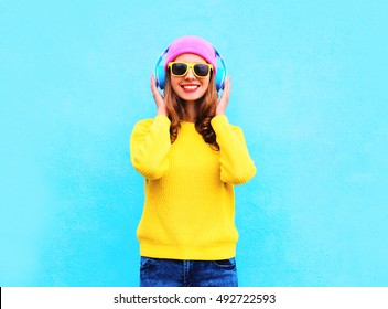 Fashion Pretty Cool Smiling Girl Listening To Music In Headphones Wearing Colorful Pink Hat, Yellow Sunglasses And Sweater Over Blue Background