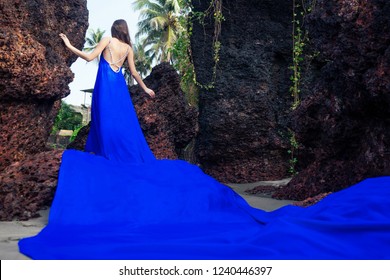 fashion pretty female model posing on a beach with rocks in a long butterfly chameleon dress waterfall of skirt plume train.sensual perfume with a beautiful and young brunette woman wave tropical sand