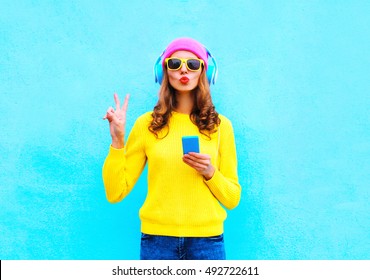 Fashion pretty sweet carefree woman listening music in headphones with smartphone wearing a colorful pink hat yellow sweater sunglasses over blue background
