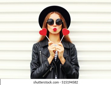 Fashion Pretty Sweet Young Woman With Red Lips Making Air Kiss With Lollipop Heart Wearing Black Hat Leather Jacket Over White Background