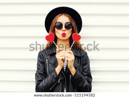 Fashion pretty sweet young woman with red lips making air kiss with lollipop heart wearing black hat leather jacket over white background