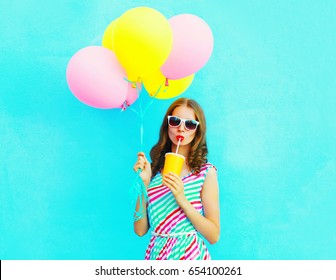 Fashion pretty woman drinks fruit juice from cup holds an air colorful balloons on a blue background