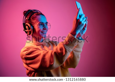 Fashion pretty woman with headphones listening to music and making selfie photo over red neon background at studio.