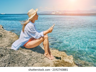 Fashion Pretty Woman Outdoors Lifestyle Watching, Reading On Tablet Ebook On The Beach In Summer Day. Wearing Wide Brimmed Hat, Sunbating With Uv Protection. Concept Of Beach Vacation.