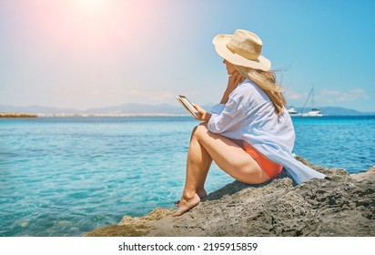 Fashion Pretty Woman Outdoors Lifestyle Watching, Reading On Tablet Ebook On The Beach In Summer Day. Wearing Wide Brimmed Hat, Sunbating With Uv Protection. Concept Of Beach Vacation.