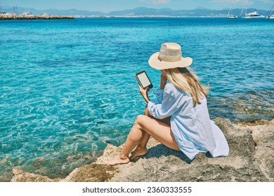 Fashion pretty woman outdoors lifestyle watching, reading on tablet ebook on the beach in summer day. Wearing wide brimmed hat, Sunbating with uv protection. Concept of beach vacation.