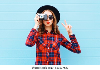 Fashion pretty young woman blowing red lips with retro camera wearing a black hat, red checkered shirt over blue background - Powered by Shutterstock