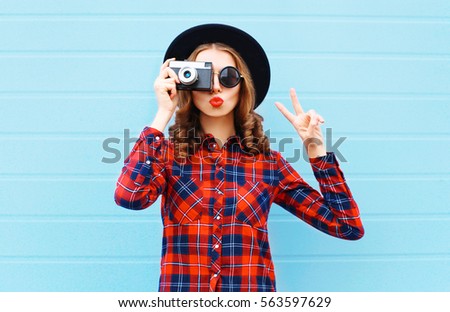 Fashion pretty young woman blowing red lips with retro camera wearing a black hat, red checkered shirt over blue background
