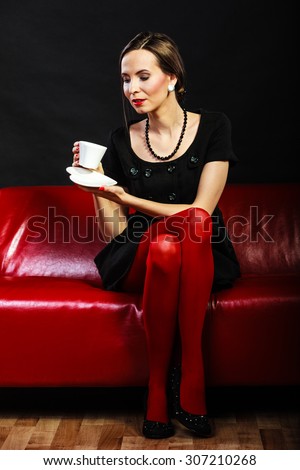 Fashion and relaxation concept. Woman retro style in full length. Elegant lady holding coffee tea cup hot drink sitting on red sofa, indoor