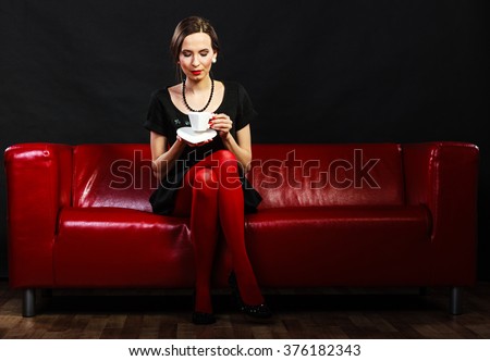 Fashion and relaxation concept. Woman retro style in full length. Elegant lady holding coffee tea cup hot drink sitting on red sofa, indoor