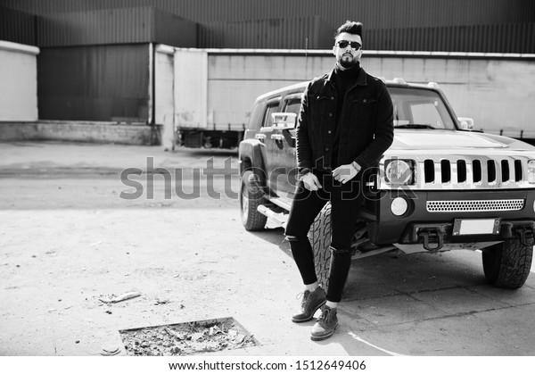 Fashion rich beard Arab man wear on\
black jeans jacket and sunglasses posed against big black suv car.\
Stylish, succesful and fashionable arabian model\
guy.