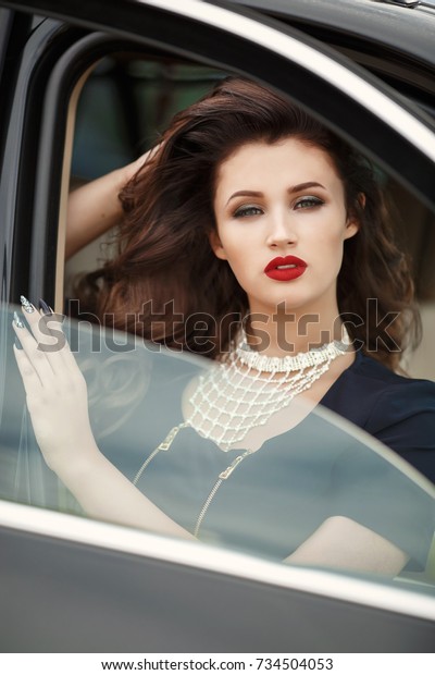 Fashion sexy woman in luxury black car posing\
outdoors. Glamour model with red lips and retro hairstyle curls in\
sport car st street. Vogue style girl in black dress with bright\
makeup and automobile.