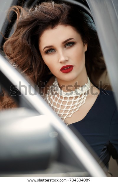 Fashion sexy woman in luxury black car posing\
outdoors. Glamour model with red lips and retro hairstyle curls in\
sport car st street. Vogue style girl in black dress with bright\
makeup and automobile.