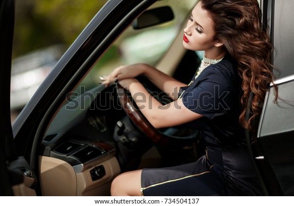 Fashion sexy woman in luxury black car posing
outdoors. Glamour model with red lips and retro hairstyle curls in
sport car st street. Vogue style girl in black dress with bright
makeup and automobile.