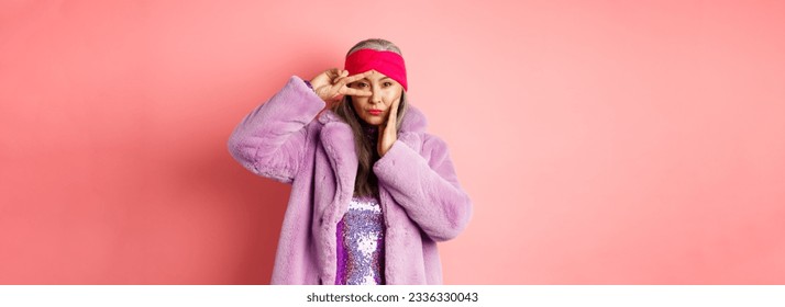 Fashion and shopping. Beautiful asian senior woman in stylish faux fur coat and headband, making peace sign on face and looking sassy and confident at camera, pink background. - Powered by Shutterstock