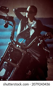 Fashion Shot. Confident Young Woman Biker In An Elegant  Evening Dress, Black Leather Jacket And Stylish Sunglasses  Poses Next To A Motorcycle In A Dark Garage.