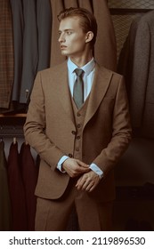 Fashion shot of a serious handsome blond man in elegant three-piece suit standing in the background of luxury classic suits in a store. Men's fashion and style.