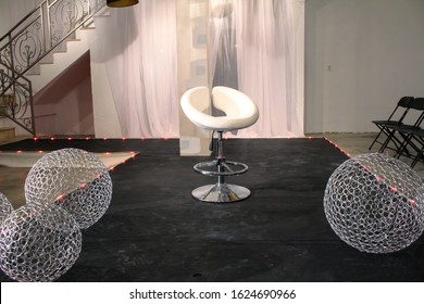 Fashion Show Runway Set Decorated With A White Seat, Led Light Strips And Silver Metal Mesh Balls On A Low Black Stage With Stairs, Folding Chairs And Sheer Drapes In The Background