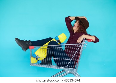 Fashion Smiling Hipster Woman Having Fun Wearing A Hat With Yellow Skateboard Sitting In The Shopping Trolley Cart Over Blue Background