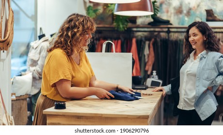 Fashion Store Owner Folding Clothes For Customer At Checkout Counter. Woman Assisting Female Shopper At Fashion Shop Checkout.