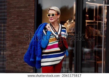 Fashion Street style autumn or spring. Model is wearing blue jacket and sweater, striped gilet, red pants and yellow sunglasses. Woman posing outside clothing store