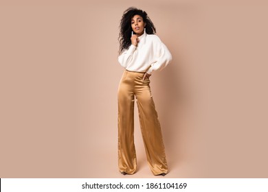 Fashion-Studio-Foto von eleganter afrikanischer Amerikanerin in beigem Hintergrund, weißer Pullover und Seidenhose. Schauen Sie sich die Kamera an. Schädellänge. 