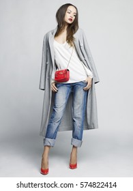 Fashion Studio Photo Of Young Stylish Woman. Grey Oversize Coat, White Shirt, Blue Jeans, Red Shoes And Handbag. Catalogue Clothes. Lookbook