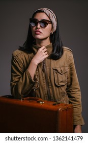 Fashion Studio Portrait Of A Beautiful, Elegant And Sophisticated Korean Asian Woman Socialite In A Brown Jacket And Sunglasses Standing Next To Her Brown Leather Suitcase. She Looks Glamorous.