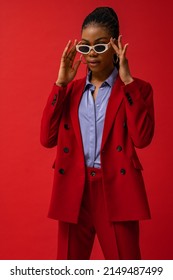 Fashion studio portrait of elegant Black woman wearing classic red suit, satin blue shirt, trendy sunglasses