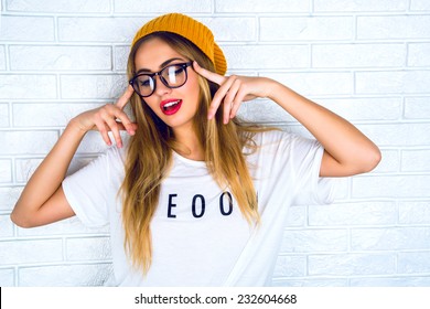 Fashion Studio Portrait Of Pretty Young Hipster Blonde Woman With Bright Sexy Make Up And Glasses , Wearing Stylish Urban T Shirt And Hat, White Urban Wall Background. 