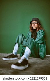 Fashion, style and clothes with a model black woman sitting on the floor against a green background. Portrait, glasses and fashionable with a young female posing to promote trendy or edgy clothing