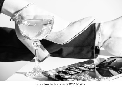 Fashion And Style Concept. Fancy Accessories, Casino Tokens, Sunglasses, Jewelry And Drink Placed On Table. Woman Legs With Black Trousers And High Heel Shoes In Background. Black And White Image