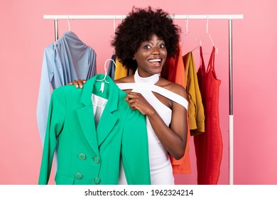 Fashion And Style. Excited Black Woman Shopping Choosing Clothes Holding Bright Jacket Smiling To Camera Posing Near Clothing Rail Over Pink Studio Background. Trendy Outfit, Shopaholism