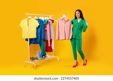 Fashion and style. Happy lady shopping and choosing clothes, holding new shirt and smiling, standing near clothing rail over yellow studio background. Shopaholism concept - Powered by Shutterstock