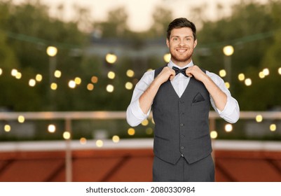 fashion, style and holidays concept - happy man in festive suit adjusting bowtie at rooftop party over lights background
