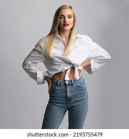 Fashion, Style And Make-up Concept. Beautiful And Sexy Long Hair Blonde Woman With Classic Outfit And Red Lipstick Studio Portrait. Slim Model Wearing Blue Jeans And White Blouse. Visible Belly