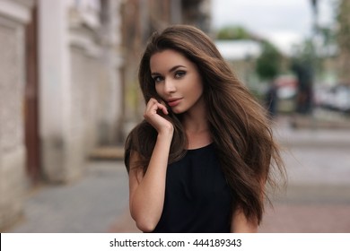 Fashion style portrait of young beautiful elegant woman in black dress walking at city streets on a windy dull day.