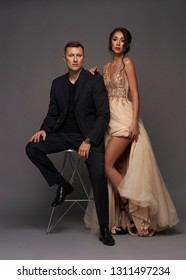 Fashion Style Studio Portrait Of Young Adult Couple. Pretty Sexy Lady In Luxury Elegant Evening Dress Standing Near Man In Black Suit Sitting On High Chair.