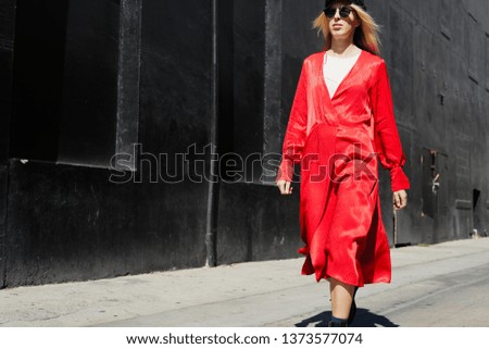 Fashion stylish woman with long blonde hair in bright red dress walking at street, black wall behind, sunny day.