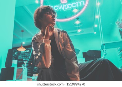 Fashion Tomboy Teen Hipster Short Hairstyle Gen Z Girl Wear Stylish Clothes Glasses Sitting On Street Near Neon Sign, Female Model Woman Look Away In City Night Light 80s 90s Style Glow, Portrait