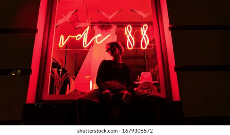Fashion Tomboy Teen Hipster Short Hairstyle Gen Z Girl Wear Stylish Clothes Glasses Sit On Red Street Window Near Neon Sign. Female Model Woman Relax In City Night Light 80s 90s Style Glow.