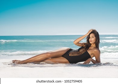 Fashion woman in black swimwear lying on tropical beach. Portrait of beautiful young woman lying on side enjoying sunbath near the sea shore. Sexy tanned girl in stylish swimsuit looking at camera.