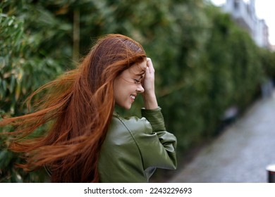 Fashion woman in the city outside posing with a smile against buildings and trees with red long flying hair in the windy weather - Powered by Shutterstock