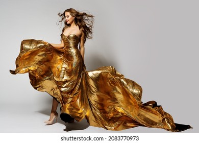 Fashion Woman Dancing in Golden Dress with Waving Hair and Flying Silk Fabric. Model in Glittering Long Evening Gown. Beauty Girl Full Length over Gray Background