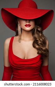 Fashion Woman in Hat hidden Face in Red Decollete Dress with Golden Earring. Elegant Lady in plunging neckline Evening Gown. Sexy Model Portrait with Glossy Red Lips over Gray Studio Background