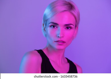 Fashion woman model posing under neon blue light headshot studio portrait. Beautiful young blond girl with natural makeup and short combed hair wearing black top looking at camera. Art design