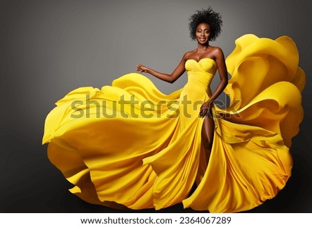 Fashion Woman in Yellow Silk Dress flowing on Wind. Dark Skinned Model with Afro Hairstyle in Long Fantasy Gown over Gray. Happy Stylish Girl dancing